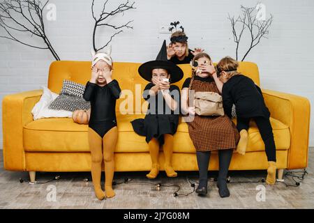 Bambini spaventati vestiti per Halloween seduti su un divano giallo, guardando il momento spaventoso in un film. Coprendo le loro bocche e gli occhi. Foto Stock