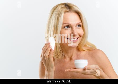 Routine di cura della pelle. Donna di media età 50s che tiene vaso con idratante anti-invecchiamento, preparando applicazione di crema sul viso, guardando via isolato su bianco. Concetto di pubblicità Foto Stock