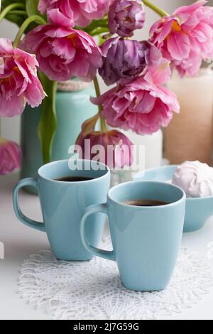 Due tazze di caffè nero e una delicata marshmallow a colazione sono accanto ad un bel bouquet di tulipani rosa in un vaso blu. Foto Stock