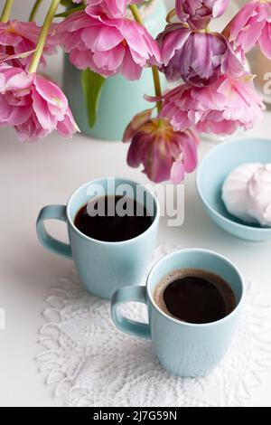 Due tazze di caffè nero e una delicata marshmallow a colazione sono accanto ad un bel bouquet di tulipani rosa in un vaso blu. Foto Stock