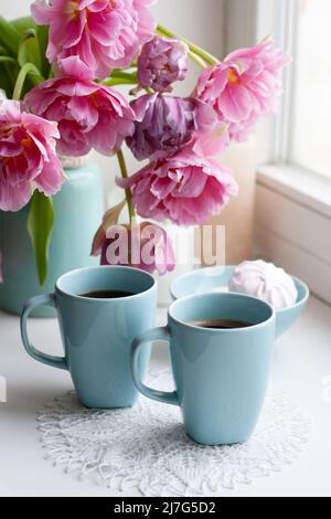 Due tazze di caffè nero e una delicata marshmallow a colazione sono accanto ad un bel bouquet di tulipani rosa in un vaso blu. Foto Stock