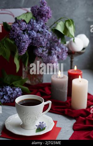 Foto d'atmosfera: Caffè nero e una tazza bianca, un bouquet di lilla, alcune grandi candele, marshmallows su sfondo grigio Foto Stock