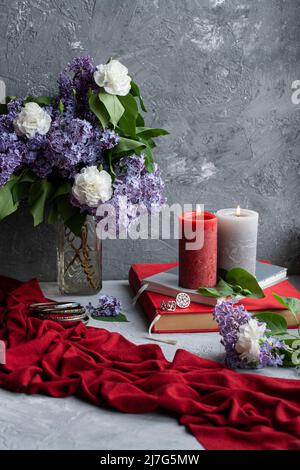 Foto d'atmosfera: Un bouquet di lilla e garofani, alcune grandi candele, libri e gioielli su sfondo grigio Foto Stock