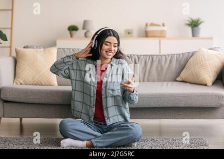 Ritratto di giovane ragazza araba allegra in cuffie che ascoltano musica su dispositivo mobile, seduto sul pavimento di casa Foto Stock