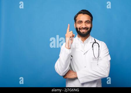 Giovane operatore medico indiano maschio che indossa abito bianco con stetoscopio su spalle in piedi isolato sfondo blu puntando dito su con successo idea, il medico trovato soluzione Foto Stock