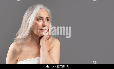 Aggraziato concetto di invecchiamento. Attraente donna anziana che tocca la pelle sul suo viso su sfondo grigio studio, panorama Foto Stock