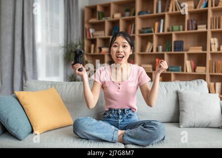 Grande gioia femminile asiatica vincitore giocare video giochi con joystick, seduta sul divano a casa, spazio copia Foto Stock