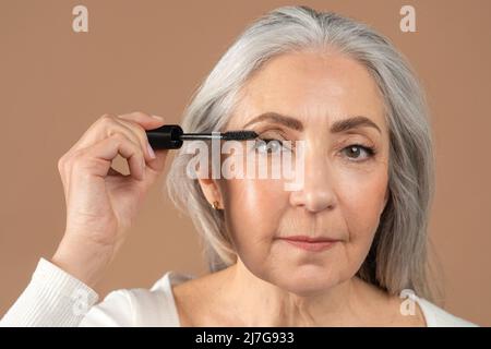 Ritratto di bellezza di affascinante donna anziana applicazione mascara sulle sue ciglia su sfondo marrone studio Foto Stock