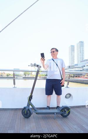 Uomo ispanico giro turistico a Puerto Madero, Buenos Aires, Argentina, con il suo scooter elettrico e prendere un selfie. Concetto: Persona anziana che utilizza techno Foto Stock