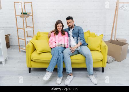 Sorridente coppia interrazziale che tiene tavolozza di colori sul divano a casa Foto Stock