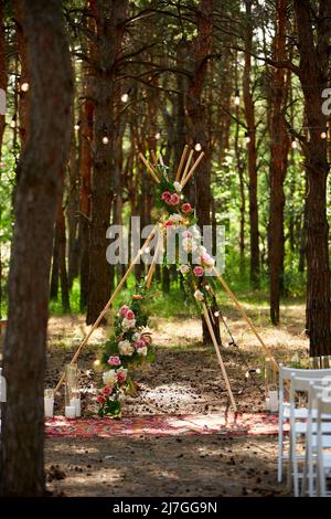 Arco di tibi bohémien fatto di aste di legno decorate con rose rosa, candele su tappeto, erba pampass, avvolto in luci fata su matrimonio all'aperto Foto Stock