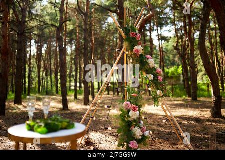 Arco di tibi bohémien fatto di aste di legno decorate con rose rosa, candele su tappeto, erba pampass, avvolto in luci fata su matrimonio all'aperto Foto Stock