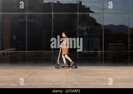 Vista laterale del giovane uomo che si trova in ufficio in una grande città con scooter elettrico per la salute ambientale e il miglioramento per evitare l'inquinamento Foto Stock