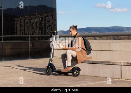 Giovane uomo moderno con pettinatura bun testando sullo smartphone mentre si sta seduto su una panca con il suo scooter elettrico sostenibile Foto Stock