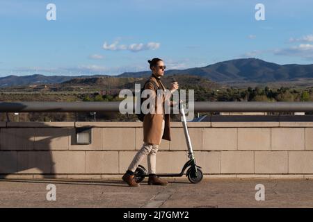 Vista laterale dell'uomo che va a lavorare su scooter elettrico con zaino e caffè a portata di mano. Paesaggio naturale sullo sfondo uno spazio di copia Foto Stock