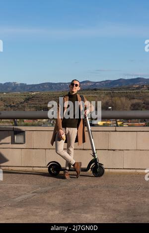 Il giovane bell'uomo sorride felicemente godendosi la giornata di sole in inverno con un caffè caldo e uno scooter elettrico per il pendolarismo Foto Stock