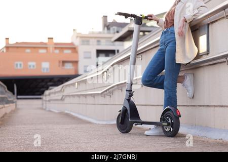 Vista panoramica di una giovane donna irriconoscibile appoggiata sulla ringhiera che tiene dietro un paesaggio urbano da scooter ad energia sostenibile Foto Stock