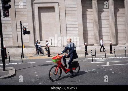Un uomo d'affari che indossa un abito corre una bicicletta a noleggio sponsorizzata da Santander lungo Threadneedle Street e oltre le alte mura della Bank of England, nella City di Londra, il quartiere finanziario della capitale, il 6th maggio 2022, a Londra, Inghilterra. Foto Stock
