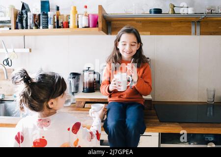 Sorridente ragazza che beve mentre sorella che tiene la tazza in cucina a casa Foto Stock