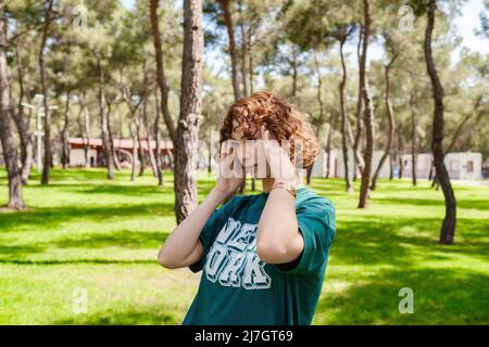 Giovane donna rossa in piedi su un parco cittadino che tocca la testa con le mani mentre ha un mal di testa sensazione di malessere, all'aperto. Signora infelice stressata Foto Stock