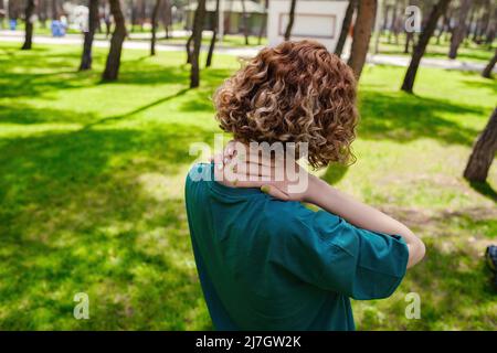 Donna redhead che indossa un tee verde dalla schiena con dolore al collo o alla spalla. Lesione o spasmo muscolare. Donna che massaggiava il collo. Assistenza sanitaria e medico con Foto Stock