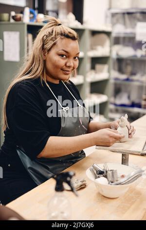 Ritratto di giovane donna sorridente con attrezzatura da lavoro e argilla modellata seduta a tavola in classe d'arte Foto Stock