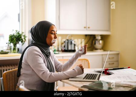 Freelance che indossa hijab facendo videochiamata attraverso il portatile seduto al tavolo in cucina Foto Stock