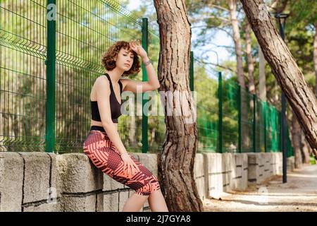 Giovane donna rossa sportiva che indossa reggiseno sportivo e leggio poggiante a parete. Donna stanca che riposa dopo l'allenamento. Modello fitness all'aperto. Correggi Foto Stock