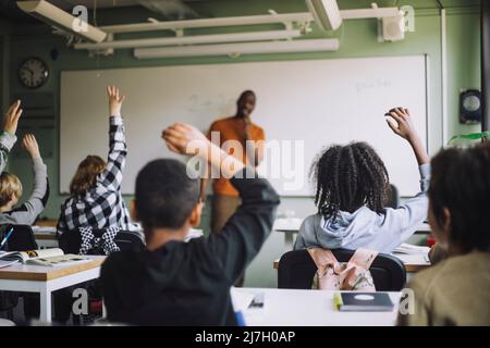 Vista posteriore degli studenti multirazziali che alzano le mani in classe durante la lezione Foto Stock