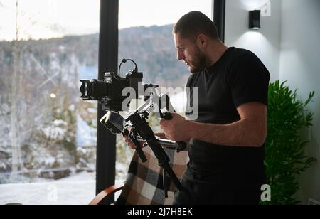 Uomo videografo che realizza riprese, utilizzando una fotocamera montata su un'attrezzatura stabilizzatore gimbal. Finestre panoramiche con paesaggio invernale sullo sfondo. Foto Stock