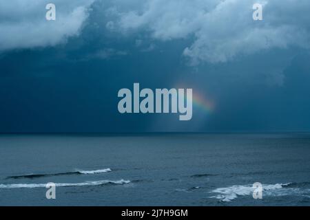 Un piccolo pezzo di arcobaleno contro un cielo di tempesta blu nero con nuvole bianche sopra e il mare in primo piano Foto Stock