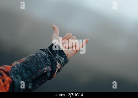 Mano maschio irriconoscibile che indossa un cappotto che rende il segno di appendere sciolto sulla cima di una montagna con picchi sullo sfondo Foto Stock