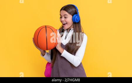 il bambino ha sorpreso le cuffie con lo zaino della scuola e la palla da basket su sfondo giallo Foto Stock