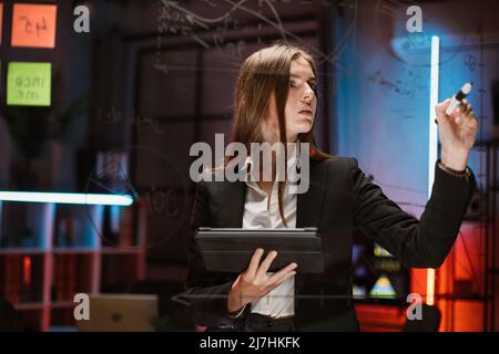 Smart donna caucasica finanza esperta in abbigliamento formale scrittura su bordo trasparente in ufficio, lavorando in serata. Dipendente aziendale che effettua report finanziari e utilizza un tablet pc Foto Stock