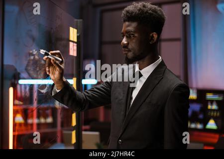 Concentrato african american maschio manager scrittura su vetro in ufficio moderno, la pianificazione avviare il progetto di business, fare note importanti e la creazione di compiti. Vista laterale ravvicinata del lavoratore in ufficio Foto Stock