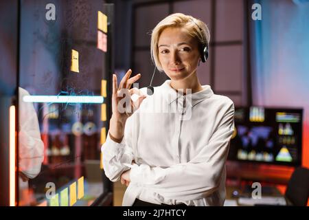 Fidata donna caucasica leader di affari in camicia bianca e cuffie, posando vicino a vetro trasparente con pennarello, guardando la fotocamera e sorridendo. Termine del progetto, concetto di pianificazione del lavoro Foto Stock