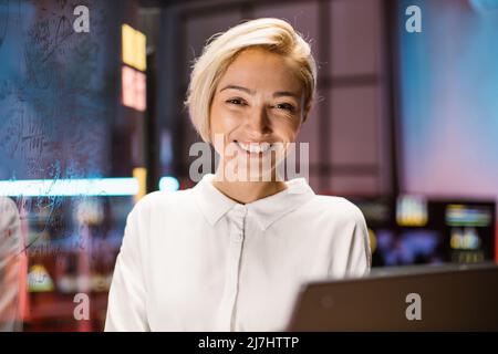 Primo piano testa di una donna caucasica piuttosto sorridente bionda in camicia bianca, in posa sulla macchina fotografica, mentre si trova in un moderno ufficio buio. Sfondo interno dell'ufficio sfocato, pannello di vetro Foto Stock