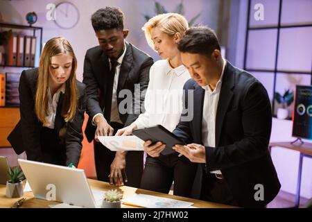 Gruppo multietnico di quattro persone motivate che lavorano insieme a un nuovo progetto, utilizzando gadget e report, condividendo idee creative. Concetto di lavoro di squadra e cooperazione Foto Stock