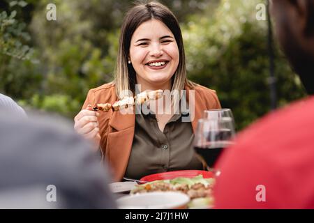 la ragazza di chubby sorride e chiacchiera con un gruppo multietnico di amici che mangiano spiedini di carne nel cortile - Una donna chubby siede al tavolo da picnic su un fresco Foto Stock