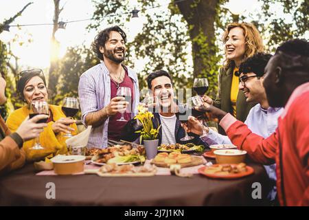 Gruppo di amici che cenano all'aperto tostando con vino rosso nel cortile - persone diverse di varie generazioni che si divertono insieme nella natura seduta Foto Stock
