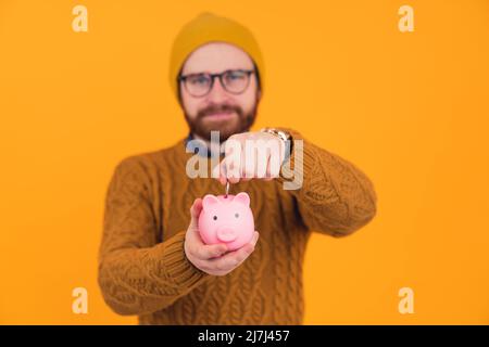 Hipster medio-vecchio ragazzo bello europeo in uno studio su sfondo arancione mettendo una moneta all'interno di un carino rosa Piggy-bank. Foto di alta qualità Foto Stock