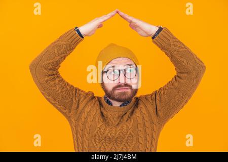 Caucasico serio preoccupato uomo rasato indossando un ponticello, una beania gialla e occhiali, tenendo le mani sopra la testa facendo un gesto sul tetto. Studio girato su sfondo arancione. Foto di alta qualità Foto Stock