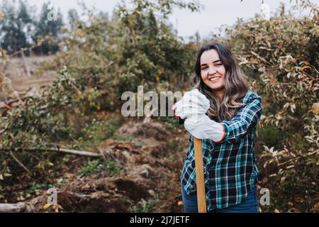 Giovane donna contadina latina sorridente con guanti da giardinaggio e appoggiata su una pala. Foto Stock