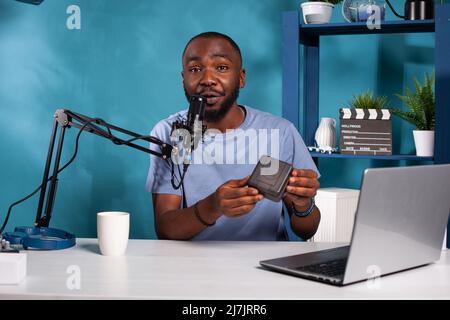 Vlogger sorridente che tiene la scatola del prodotto facendo la revisione in linea in studio di vlogging con messa a punto dal vivo professionale. Influencer che ospita prodotti in omaggio online per i fan mentre parla nel microfono. Foto Stock