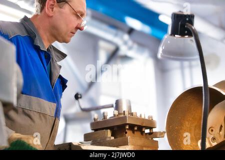 Un anziano tornitore di 50-55 anni in tute lavora al tornio in produzione. Lavorazione di prodotti metallici. Ritratto reale di lavoratore caucasico. Background di produzione. Foto Stock