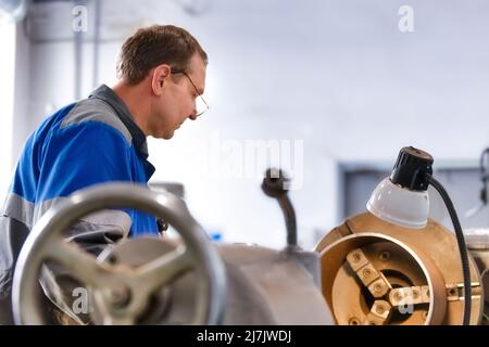 Un anziano tornitore di 50-55 anni in tute lavora al tornio in produzione. Lavorazione di prodotti metallici. Ritratto reale di lavoratore caucasico. Background di produzione. Foto Stock