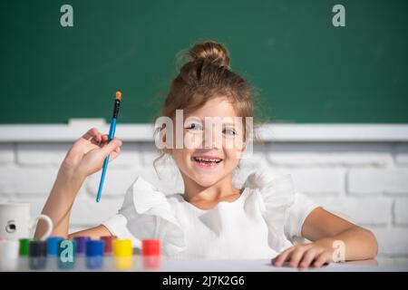 Bambina attira in classe seduto a un tavolo, divertendosi a scuola sfondo lavagna. Scuola di pittura, arte del disegno Foto Stock