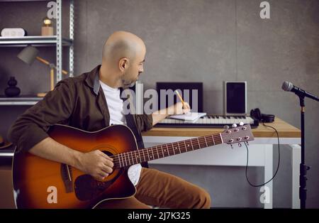 Un compositore di musica maschile di talento scrive testi e note in un notebook mentre suona la chitarra. Foto Stock