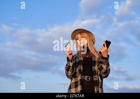 Happy hipster donna turistica facendo shopping online in background di cielo nuvoloso blu. Giovane bionda caucasica in cappello con smartphone e carta di credito. Immagine di alta qualità Foto Stock