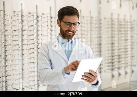 Un oftalmologo sorridente che lavora sul tablet digitale davanti alla vetrina con occhiali Foto Stock